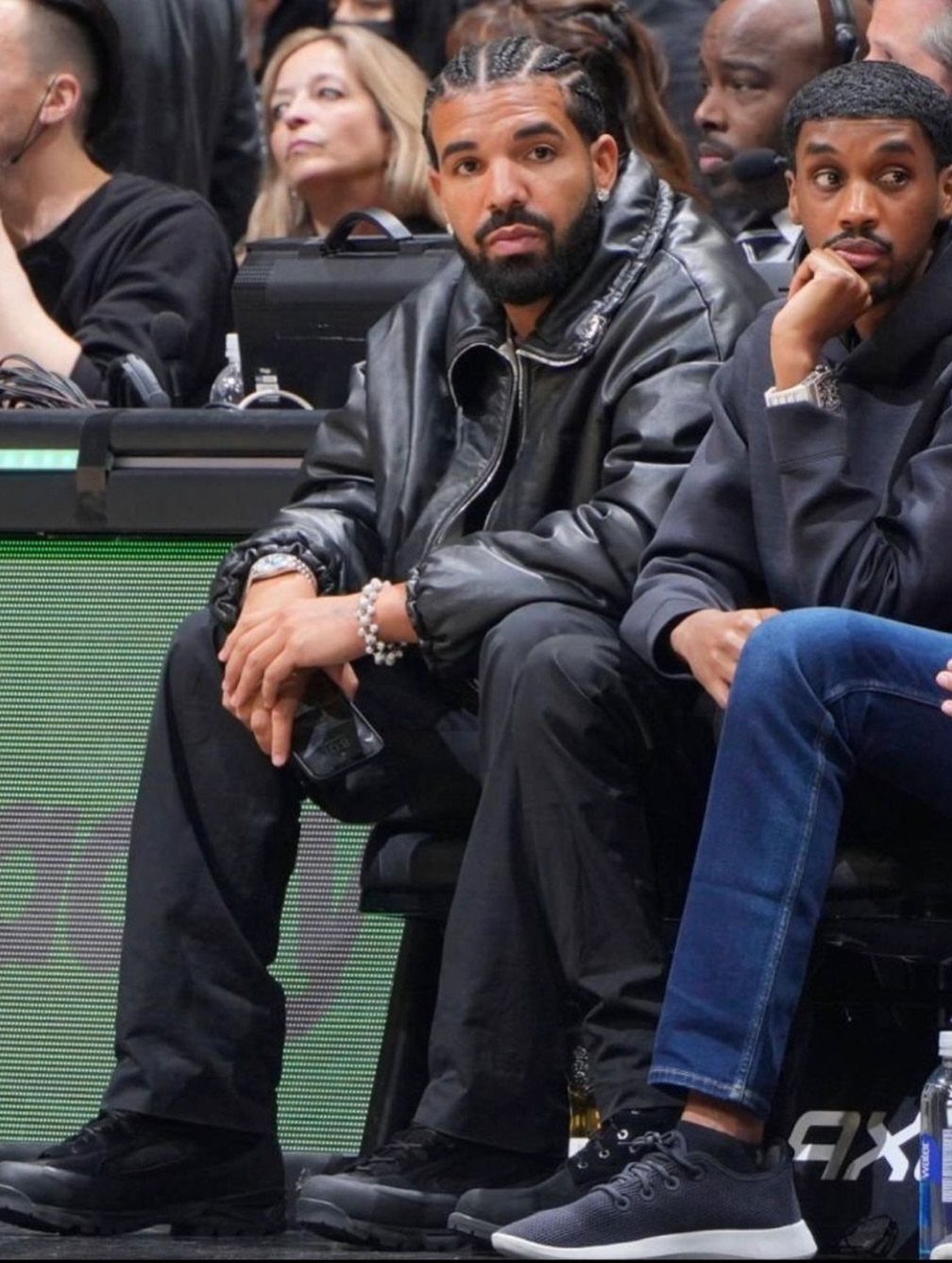 Man in an all black outfit including a leather jacket and black trousers sitting in stadium seating.