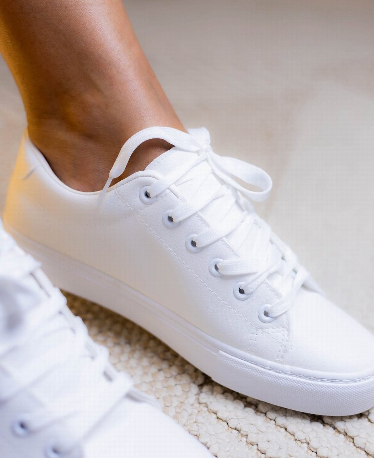 White minimalist sneakers with matching laces worn on feet over a textured rug.