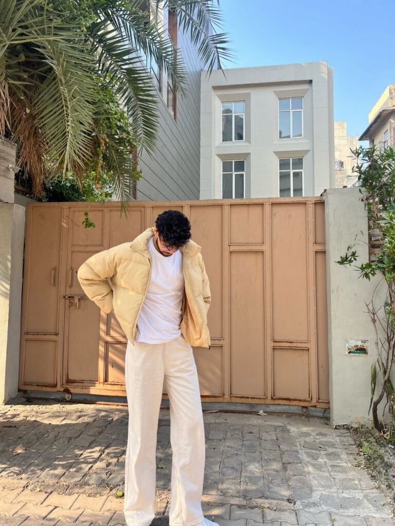 Man wearing a beige puffer jacket, white t-shirt, and cream trousers standing by a tan gate.
