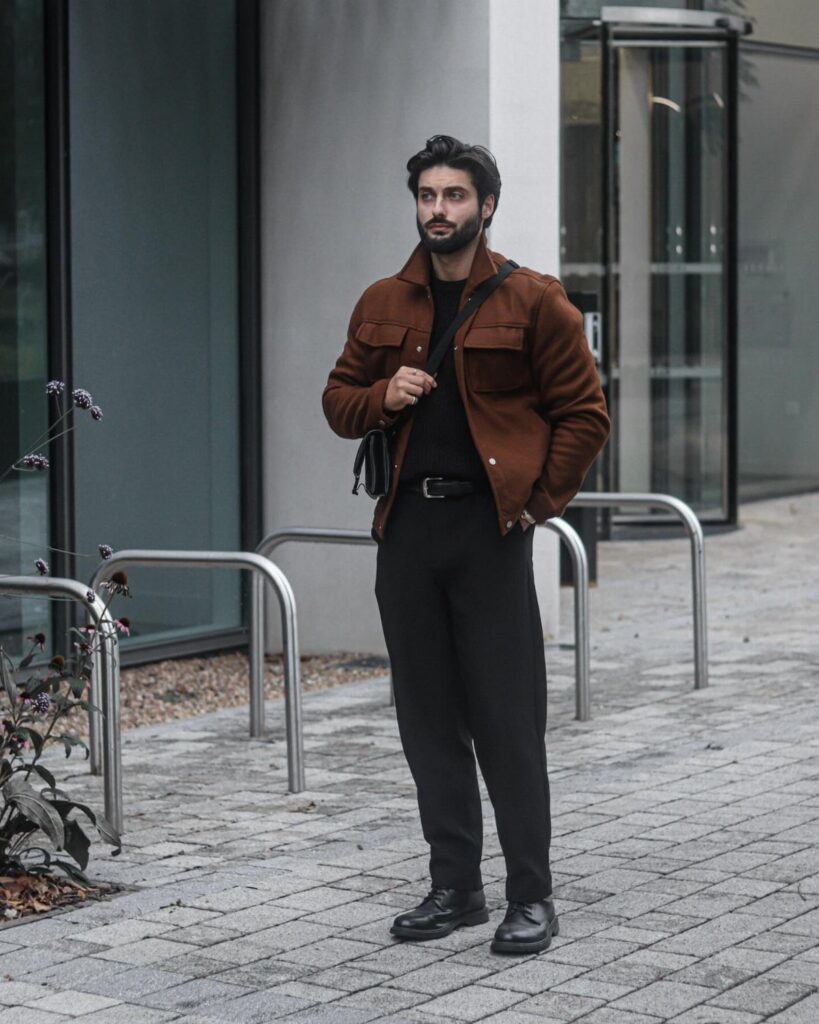 A man posing in a brown jacket, black shirt, and black trousers outdoors.