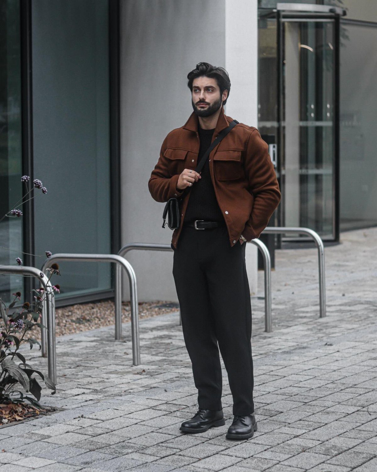 A man posing in a brown jacket, black shirt, and black trousers outdoors.