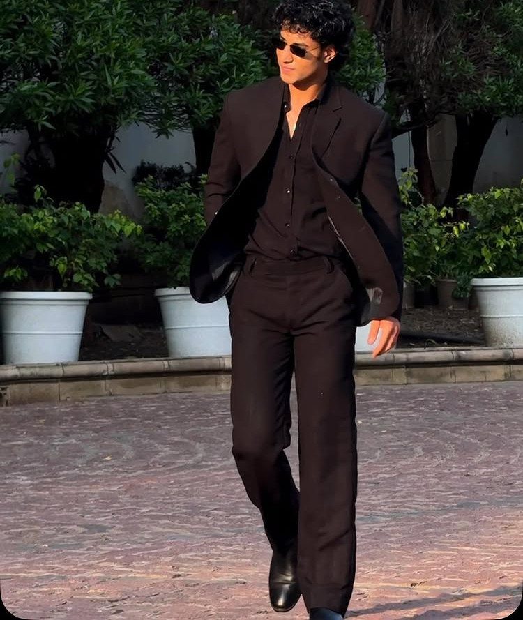 A man in a full black suit and sunglasses walking through an outdoor courtyard.
