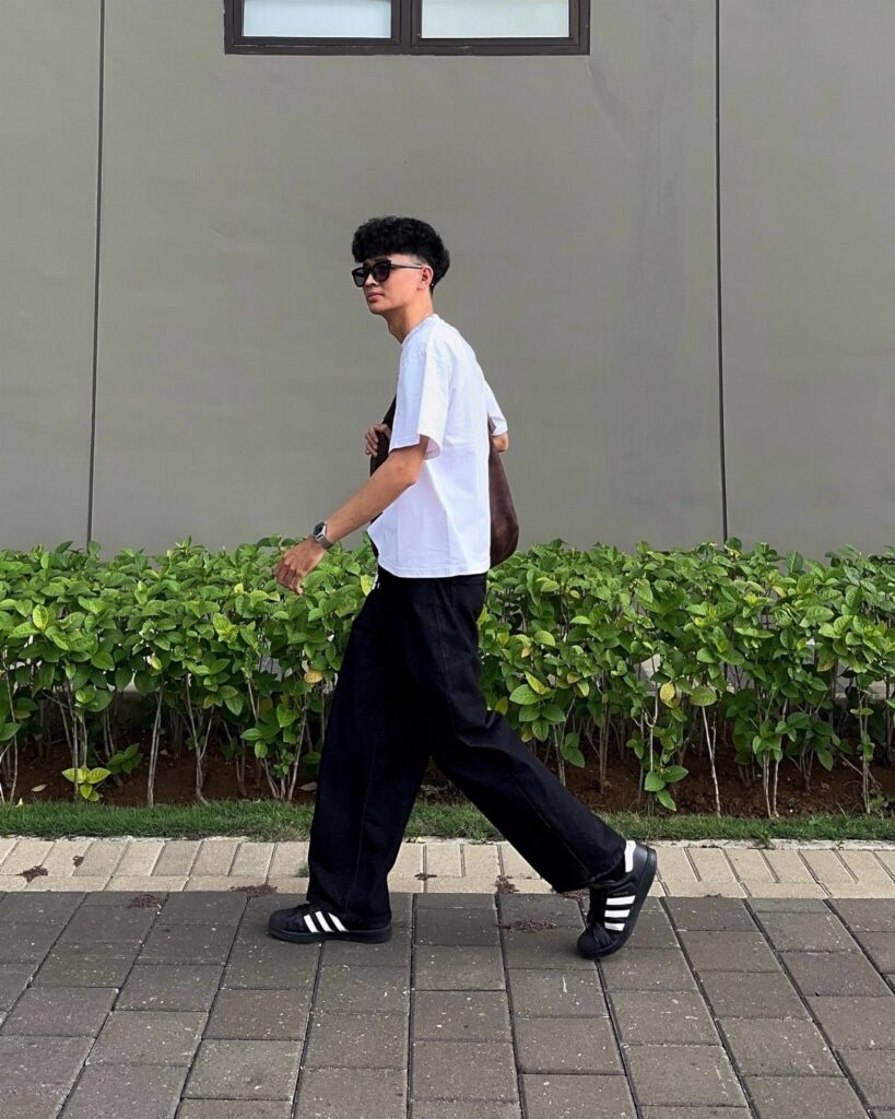 A man in a white t-shirt and black jeans walking past a green hedge.