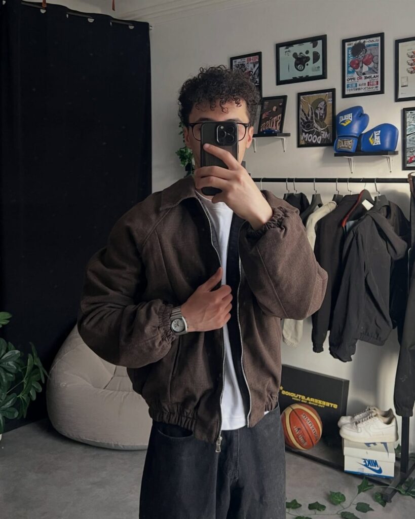 Man wearing a brown jacket, white t-shirt, and dark jeans posing for a mirror selfie.