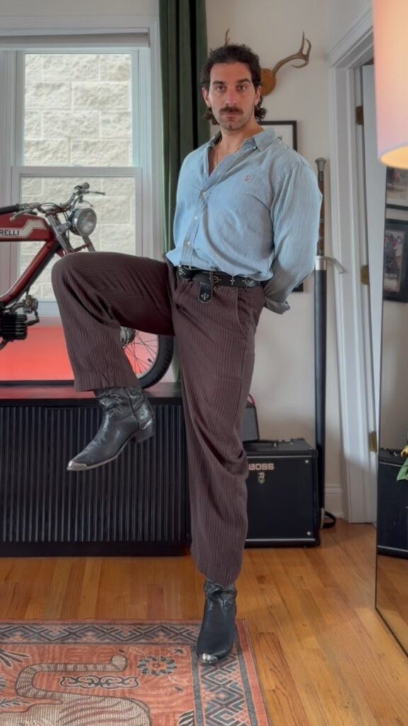 Man posing in a light blue chambray shirt and brown corduroy pants with black boots.