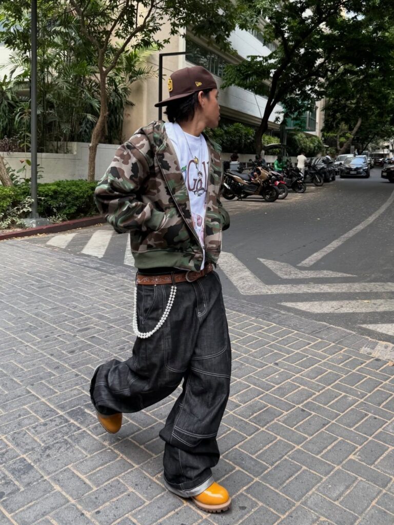 A person walking down a street wearing a camouflage jacket and baggy dark denim jeans.