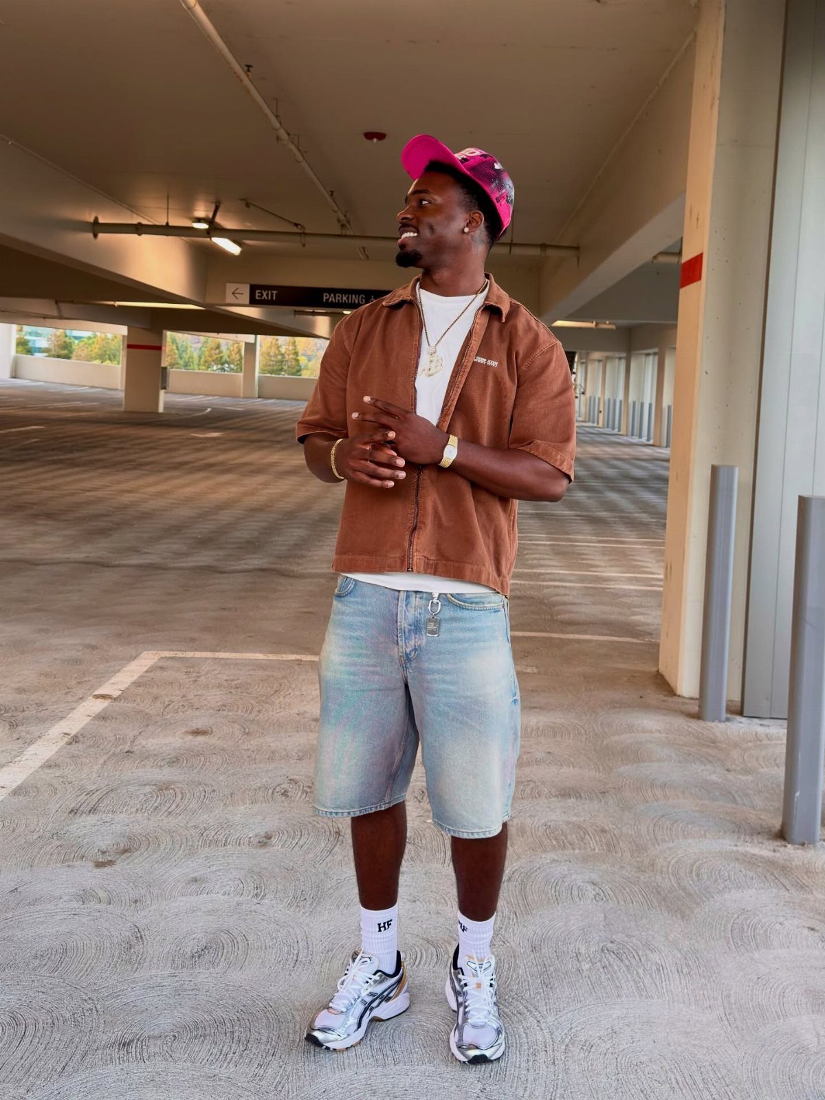 A man in a brown shirt and denim shorts wearing white chunky sneakers in a parking garage.