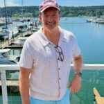 A man in a white polo shirt and light blue shorts smiling at a marina.