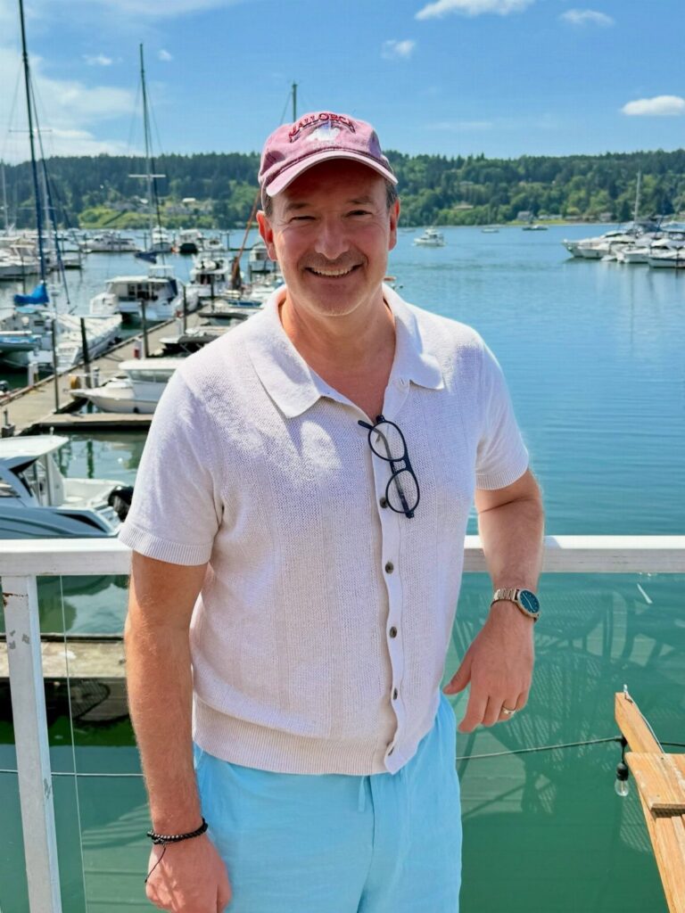 A man in a white polo shirt and light blue shorts smiling at a marina.