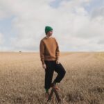 A man wearing a brown sweater, black pants, and green beanie stands in a wheat field.