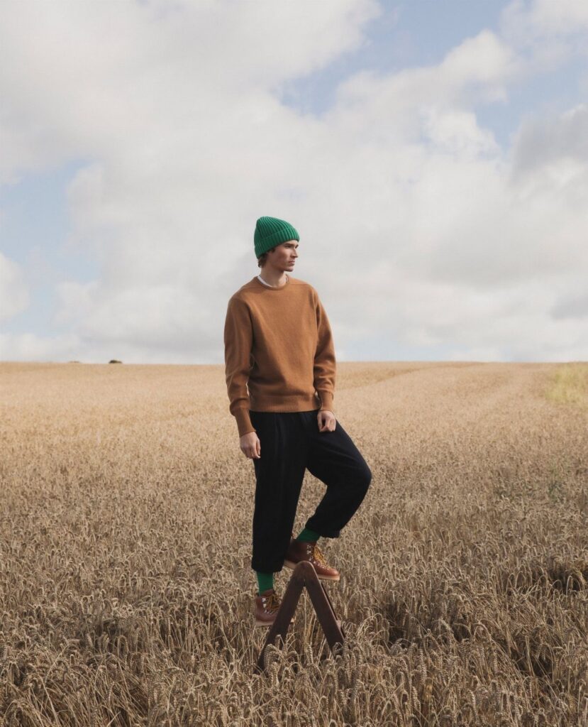 A man wearing a brown sweater, black pants, and green beanie stands in a wheat field.