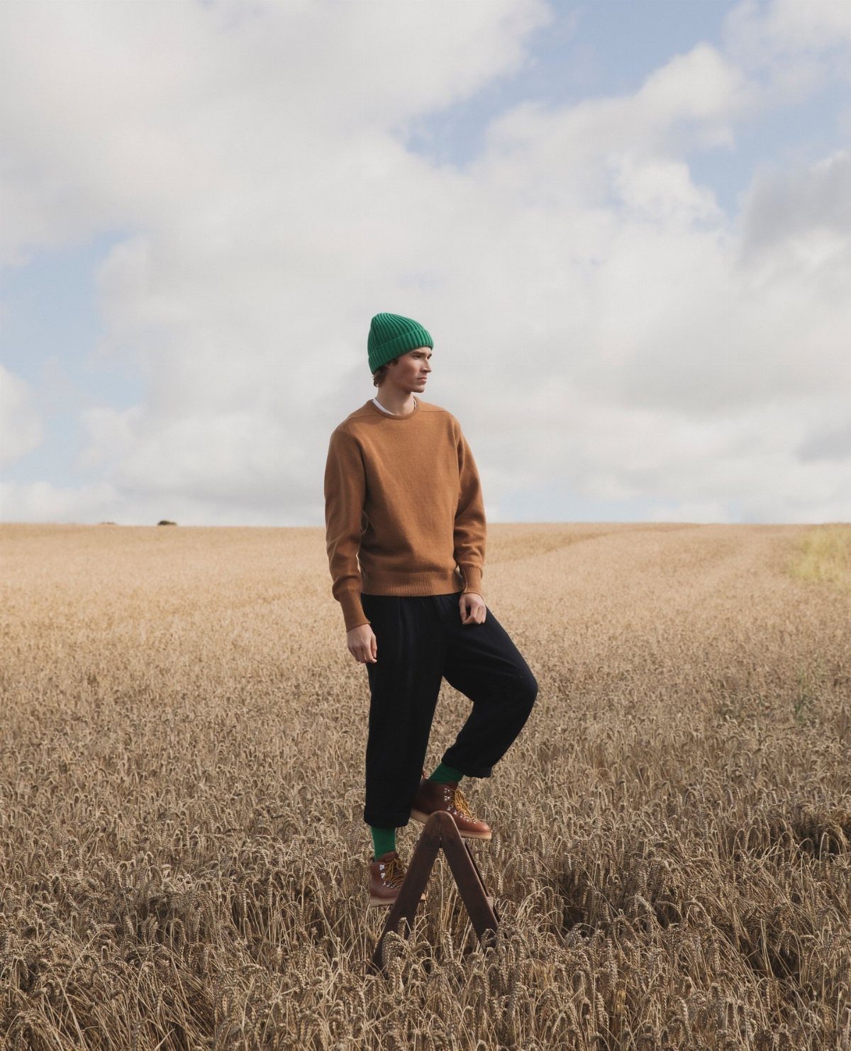 A man wearing a brown sweater, black pants, and green beanie stands in a wheat field.