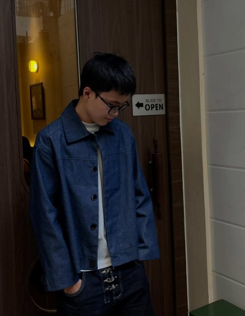 Young person wearing a blue denim jacket over a white t-shirt and dark jeans.
