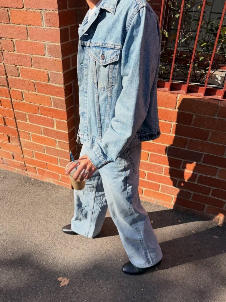 Man walking in a light blue denim jacket and matching denim jeans by a brick wall.