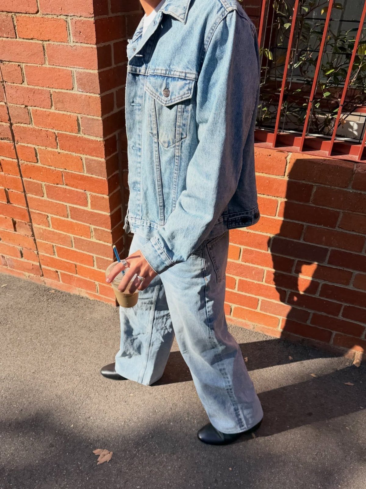 Man walking in a light blue denim jacket and matching denim jeans by a brick wall.
