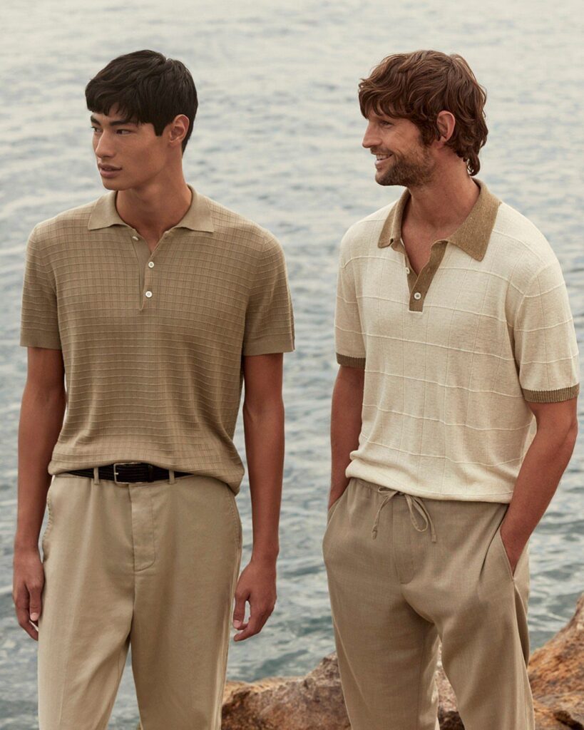 Two men wearing neutral colored polo shirts and relaxed trousers standing by the ocean.