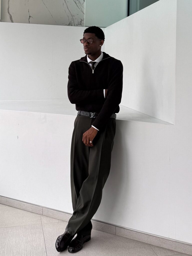 A man in smart-casual attire leaning against a white wall in a minimalist environment.