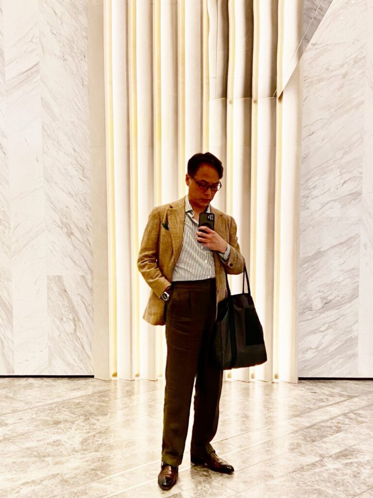 A man in a tan blazer, striped shirt, and brown trousers standing in a marble hall.