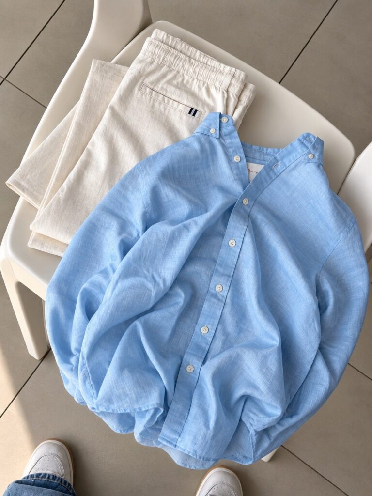 A light blue linen button-up shirt and cream trousers laid on a white chair.