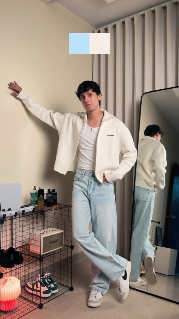 A young man posing in light blue jeans, a white jacket, and white sneakers.