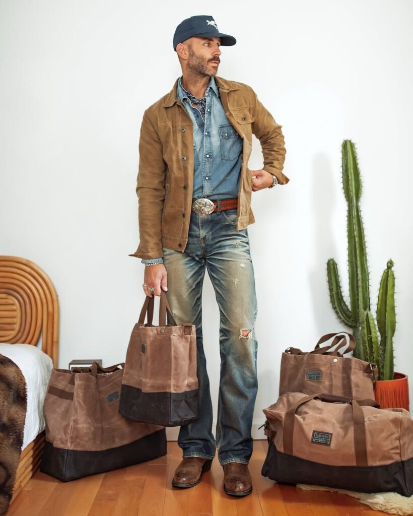 Man in denim and suede jacket wearing brown leather western boots standing near canvas bags.