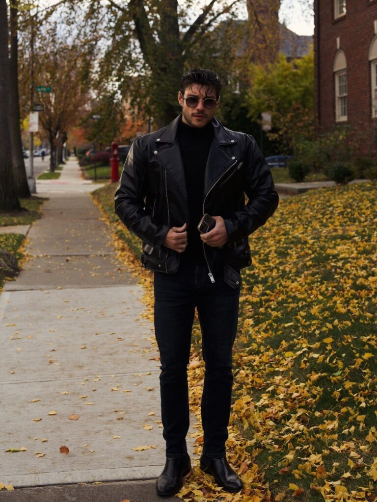 Man wearing a black leather jacket, black turtleneck, dark jeans, and sunglasses standing on a leaf-covered sidewalk.
