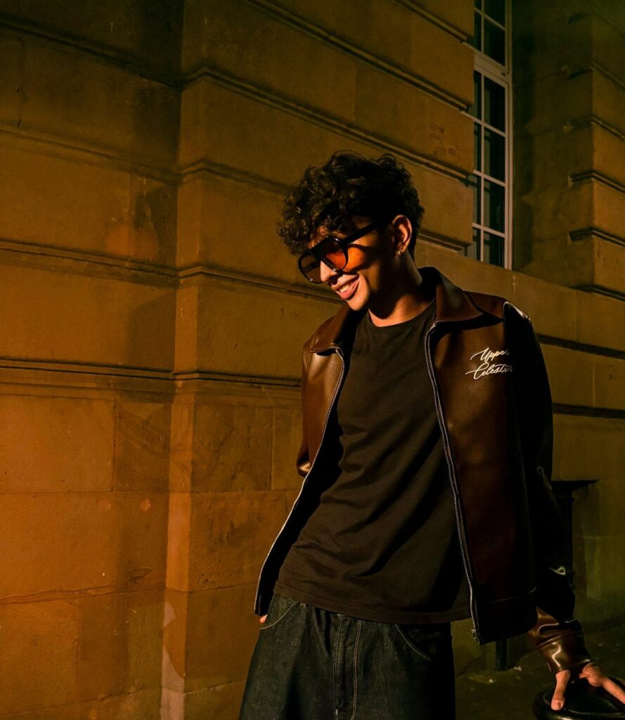 A young man wearing a dark brown leather jacket and glasses smiling against a stone wall.