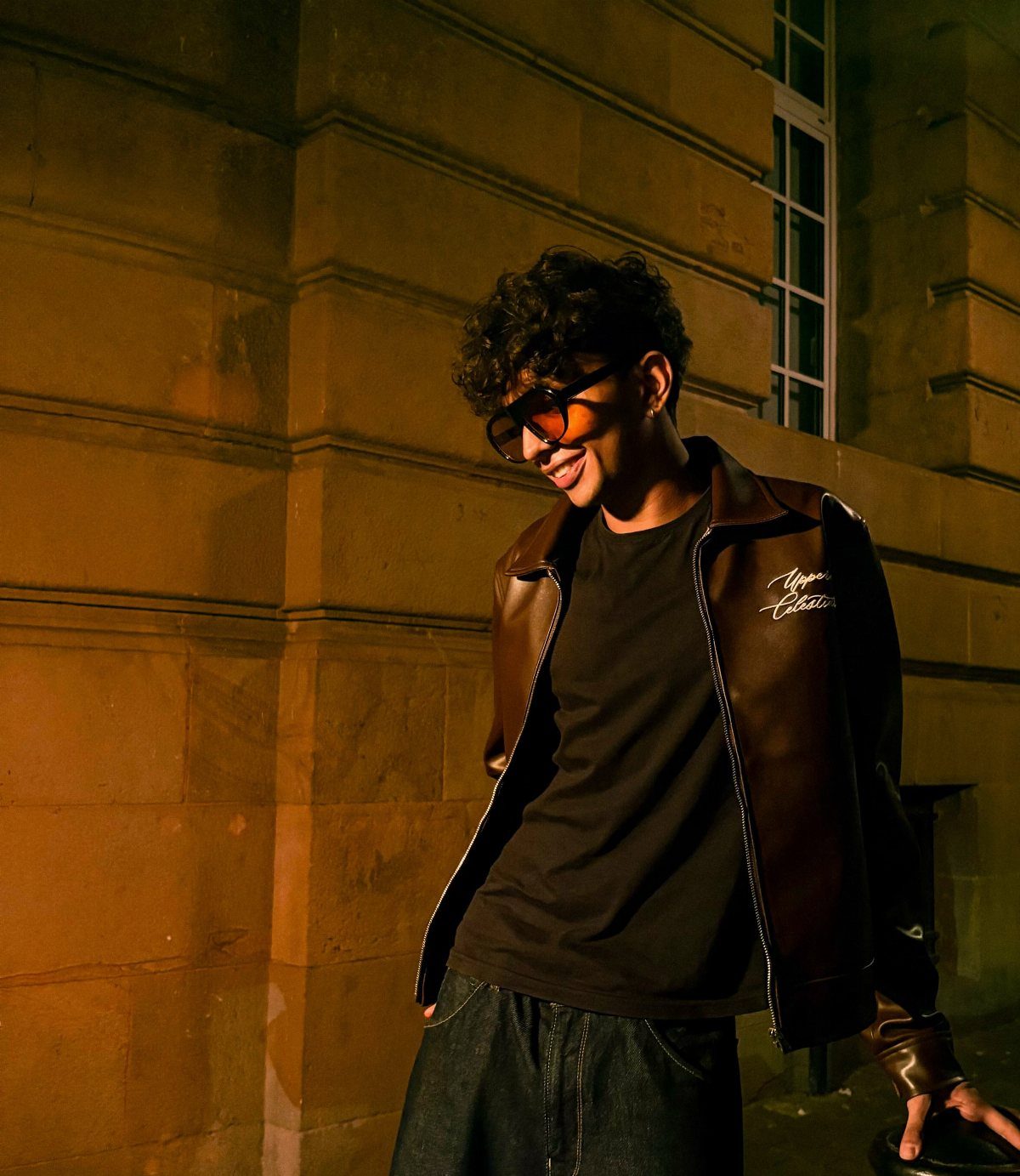 A young man wearing a dark brown leather jacket and glasses smiling against a stone wall.