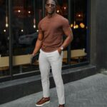A man in a brown henley, white pants, and brown shoes posing outdoors.