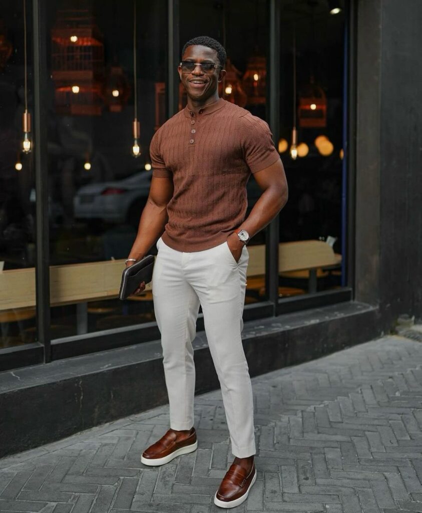 A man in a brown henley, white pants, and brown shoes posing outdoors.