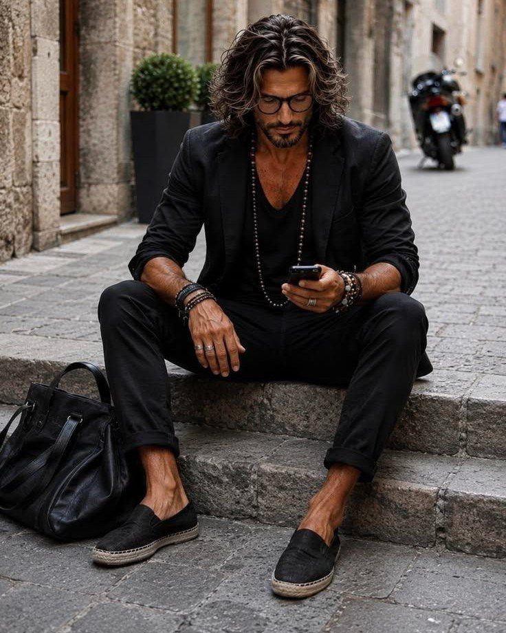 Man in all black clothing sitting on stone steps wearing a blazer, trousers, and loafers.