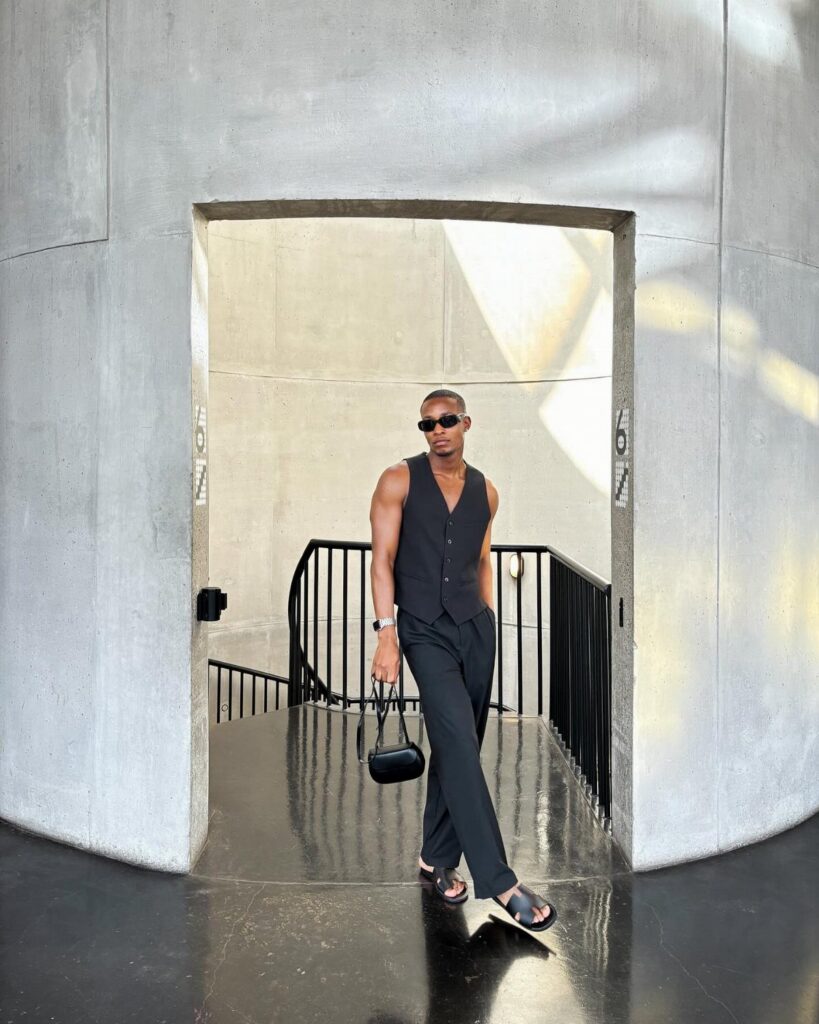 A man in a black vest and trousers walking through a concrete archway while wearing sunglasses.