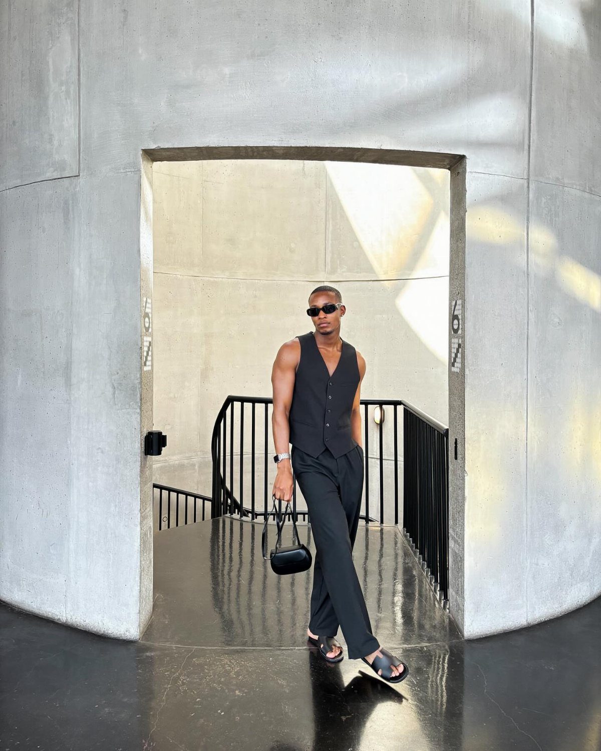 A man in a black vest and trousers walking through a concrete archway while wearing sunglasses.
