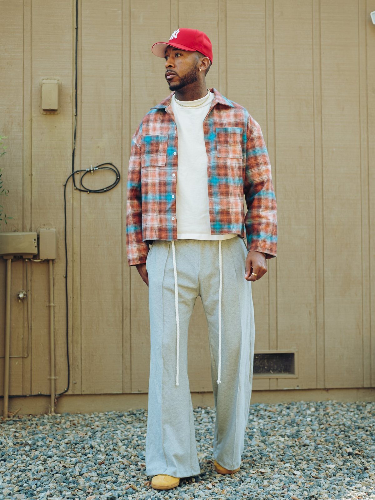 Man wearing a plaid flannel shirt, white t-shirt, and light grey sweatpants standing outdoors.