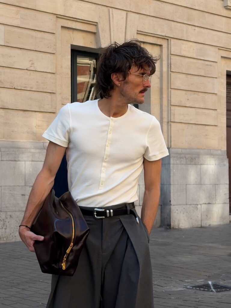 Man in a white henley shirt and dark trousers carrying a large leather bag outdoors.