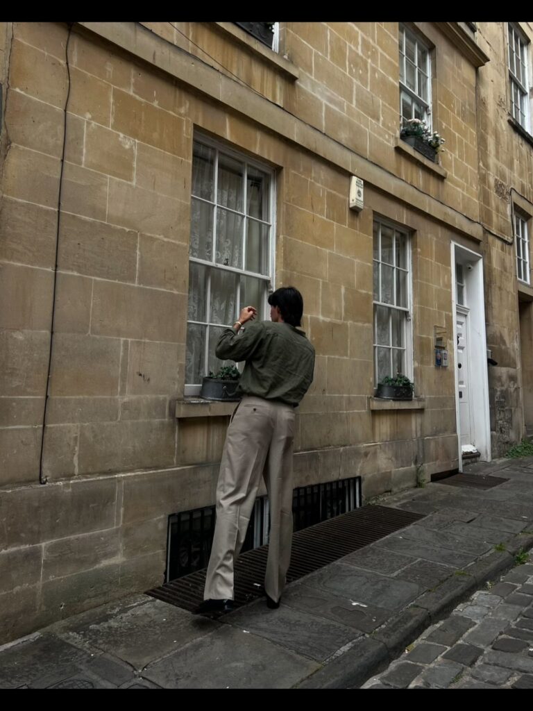 Man in olive shirt and beige trousers looking into a window of a stone building.
