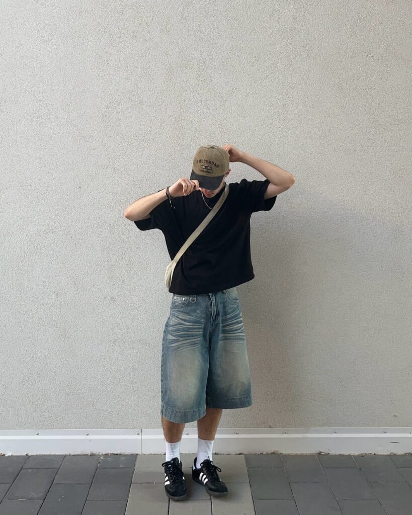 A person wearing a black t-shirt, baggy denim shorts, and a tan baseball cap against a grey wall.