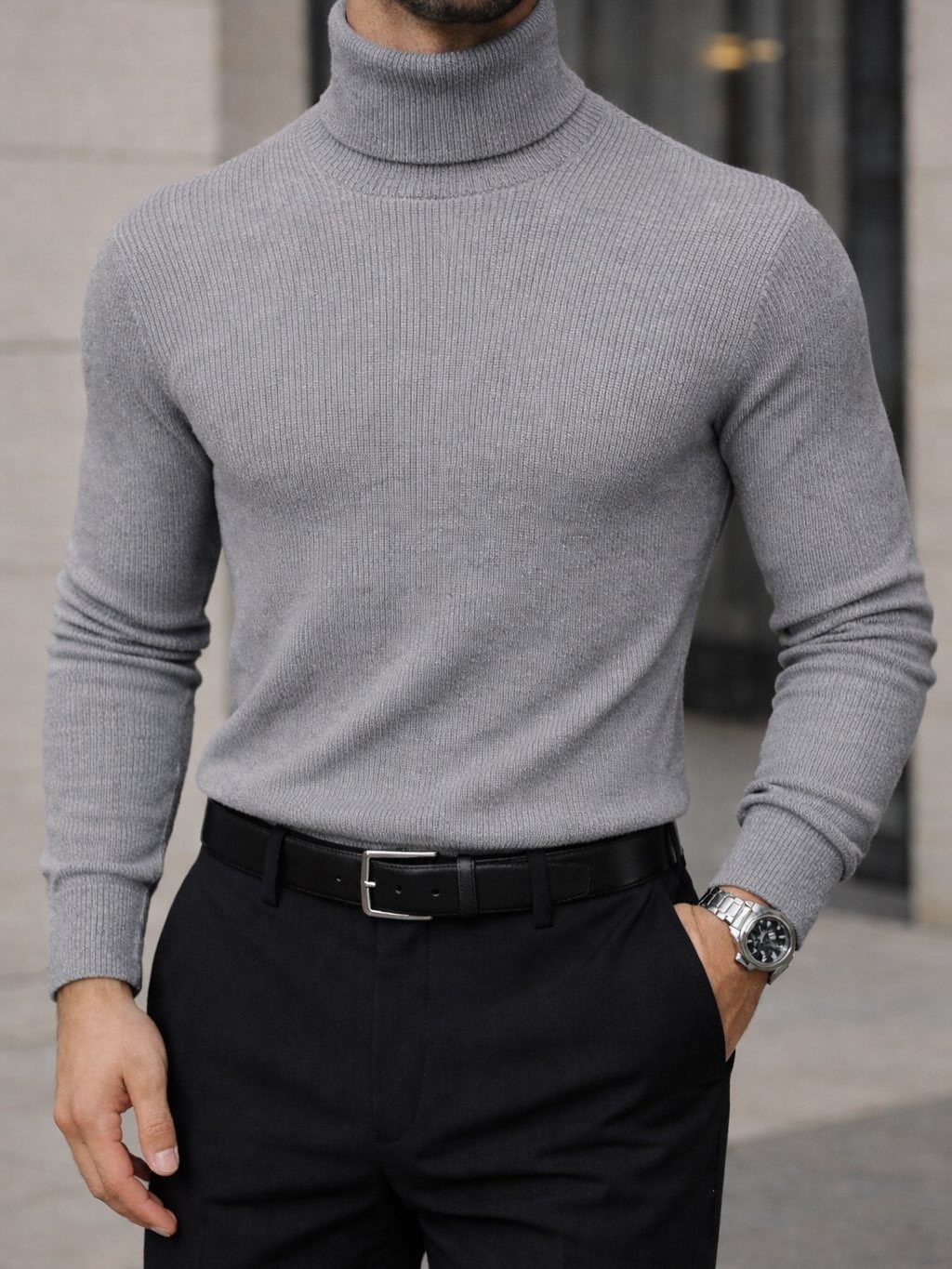 A man wearing a grey ribbed turtleneck sweater and black trousers with a silver watch.