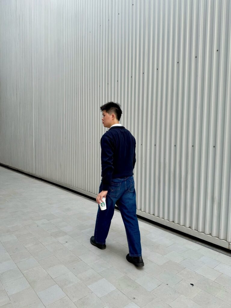 A young man in a navy shirt and blue jeans walking past a corrugated wall.