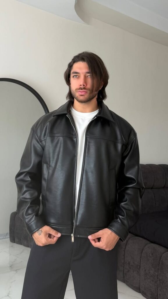 Man wearing a black leather jacket, white t-shirt, and black trousers standing indoors.
