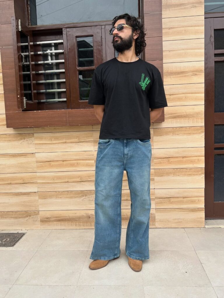 Man wearing a black graphic t-shirt and blue wide-leg jeans standing against a wooden wall.