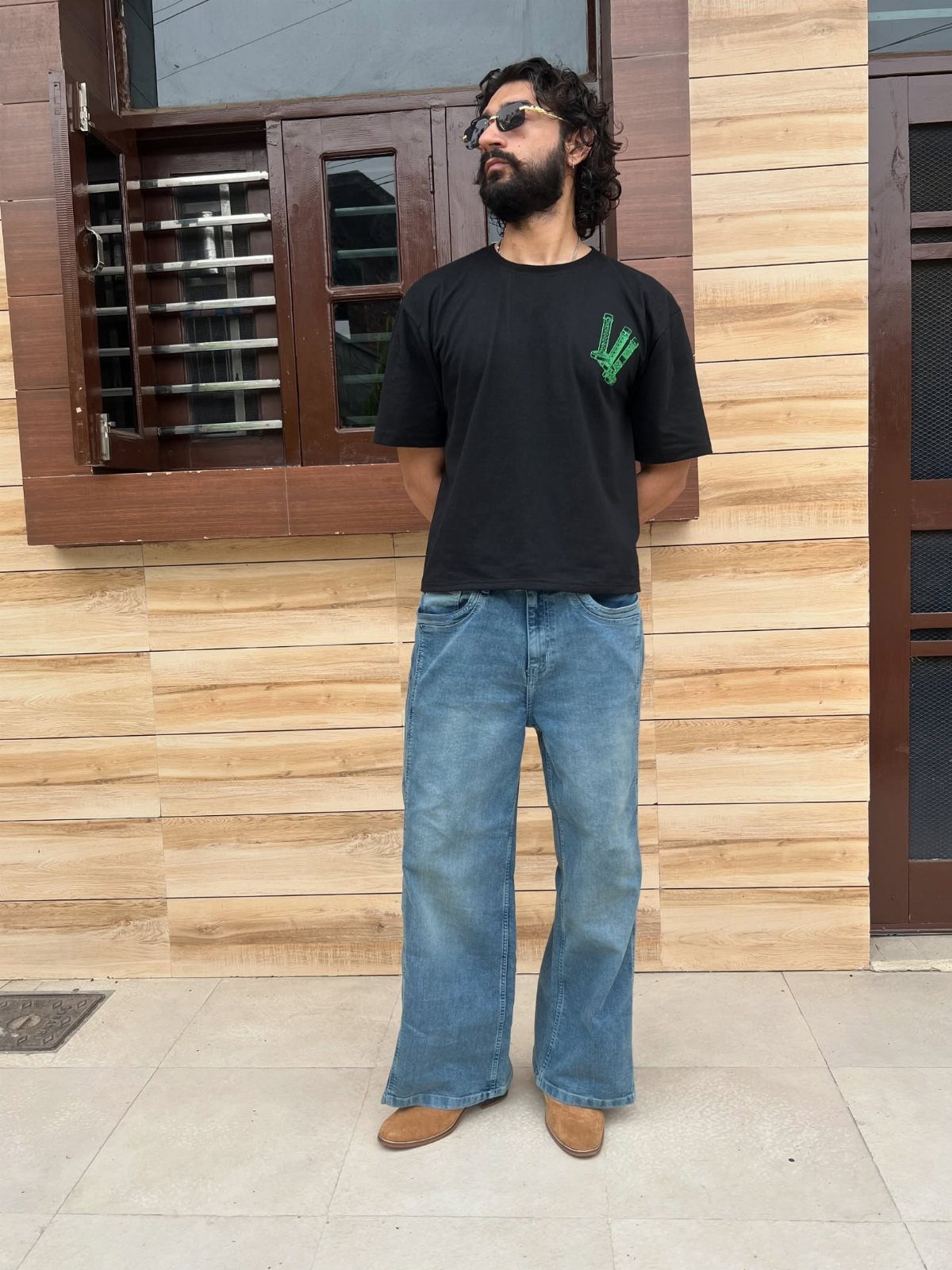Man wearing a black graphic t-shirt and blue wide-leg jeans standing against a wooden wall.