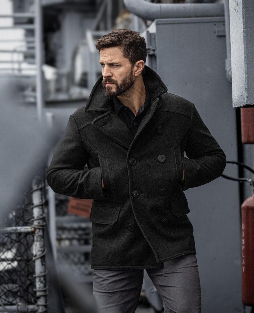 Man wearing a dark charcoal pea coat and grey trousers standing in an industrial setting.
