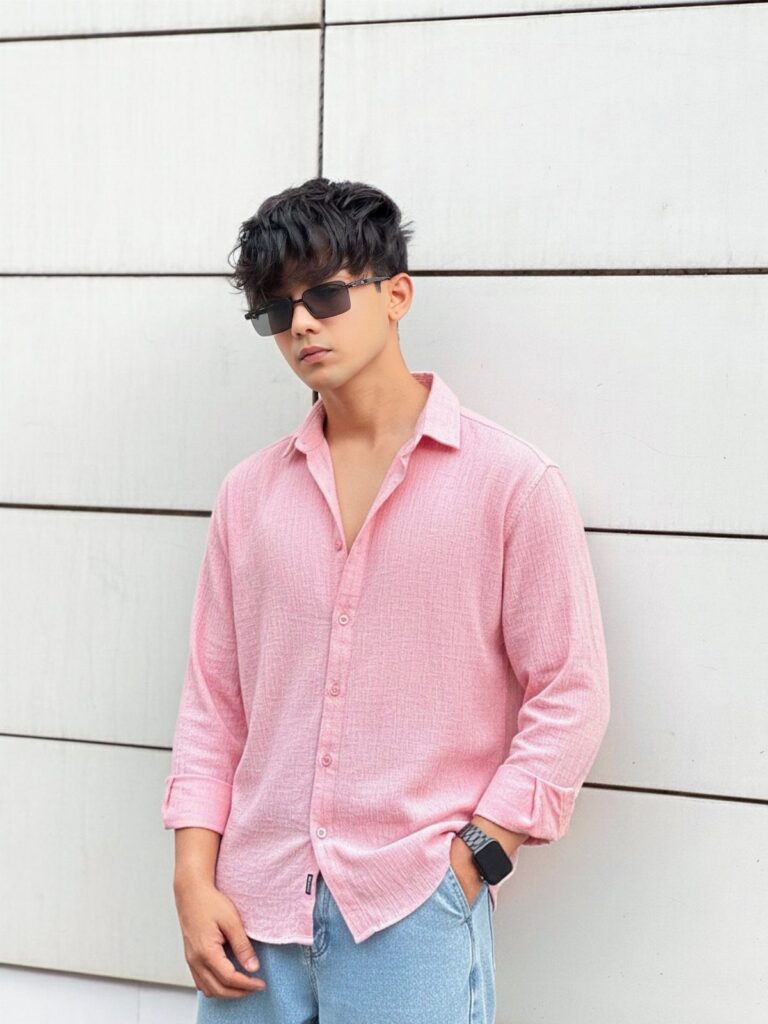 A man in a pink linen shirt and blue jeans posing against a white wall.