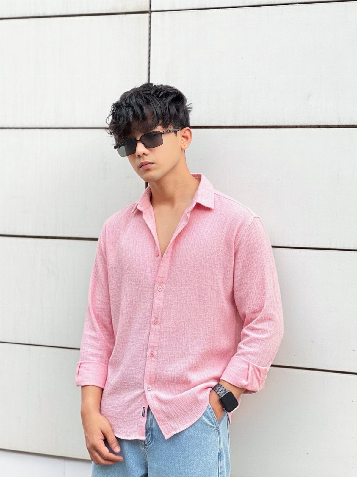A man in a pink linen shirt and blue jeans posing against a white wall.