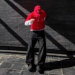 Man wearing a red baseball cap, red shirt, and black trousers standing against a brick wall.