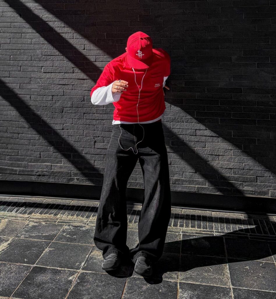 Man wearing a red baseball cap, red shirt, and black trousers standing against a brick wall.