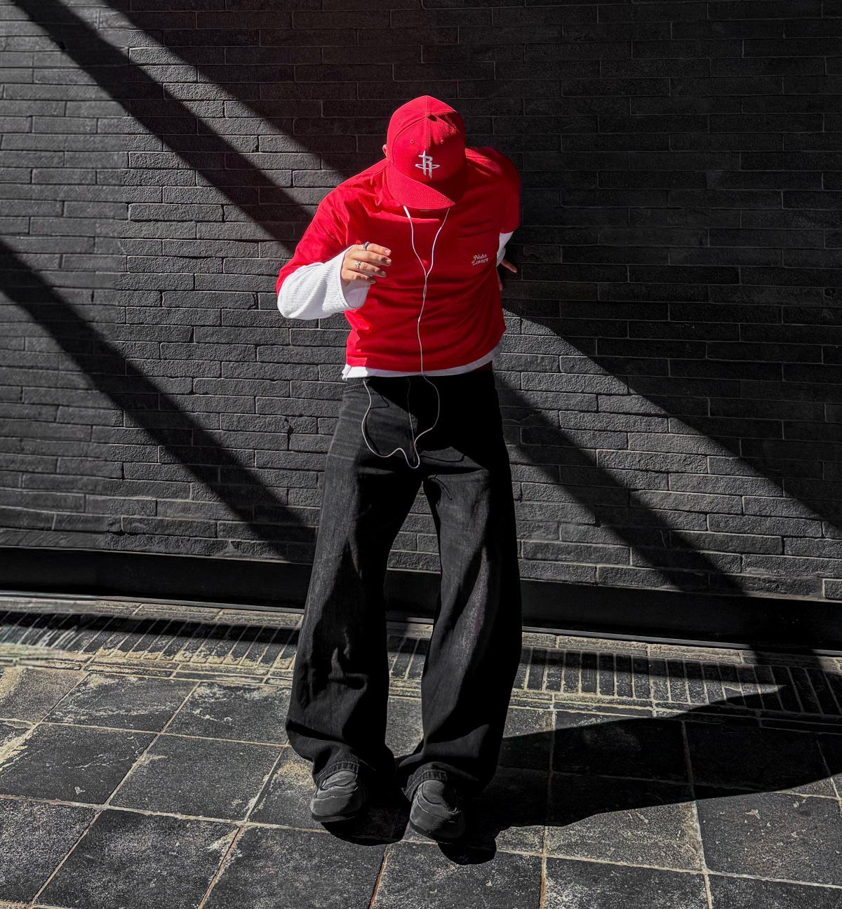 Man wearing a red baseball cap, red shirt, and black trousers standing against a brick wall.