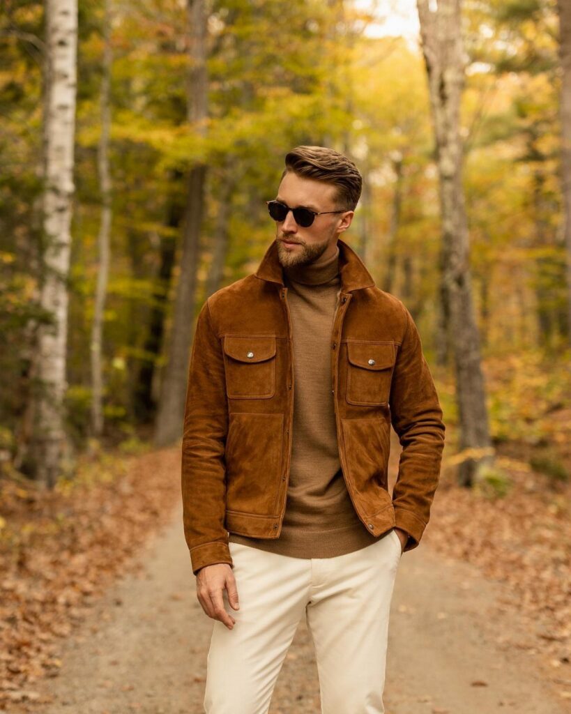Man in a brown suede jacket and turtleneck sweater standing on a forest path during autumn.