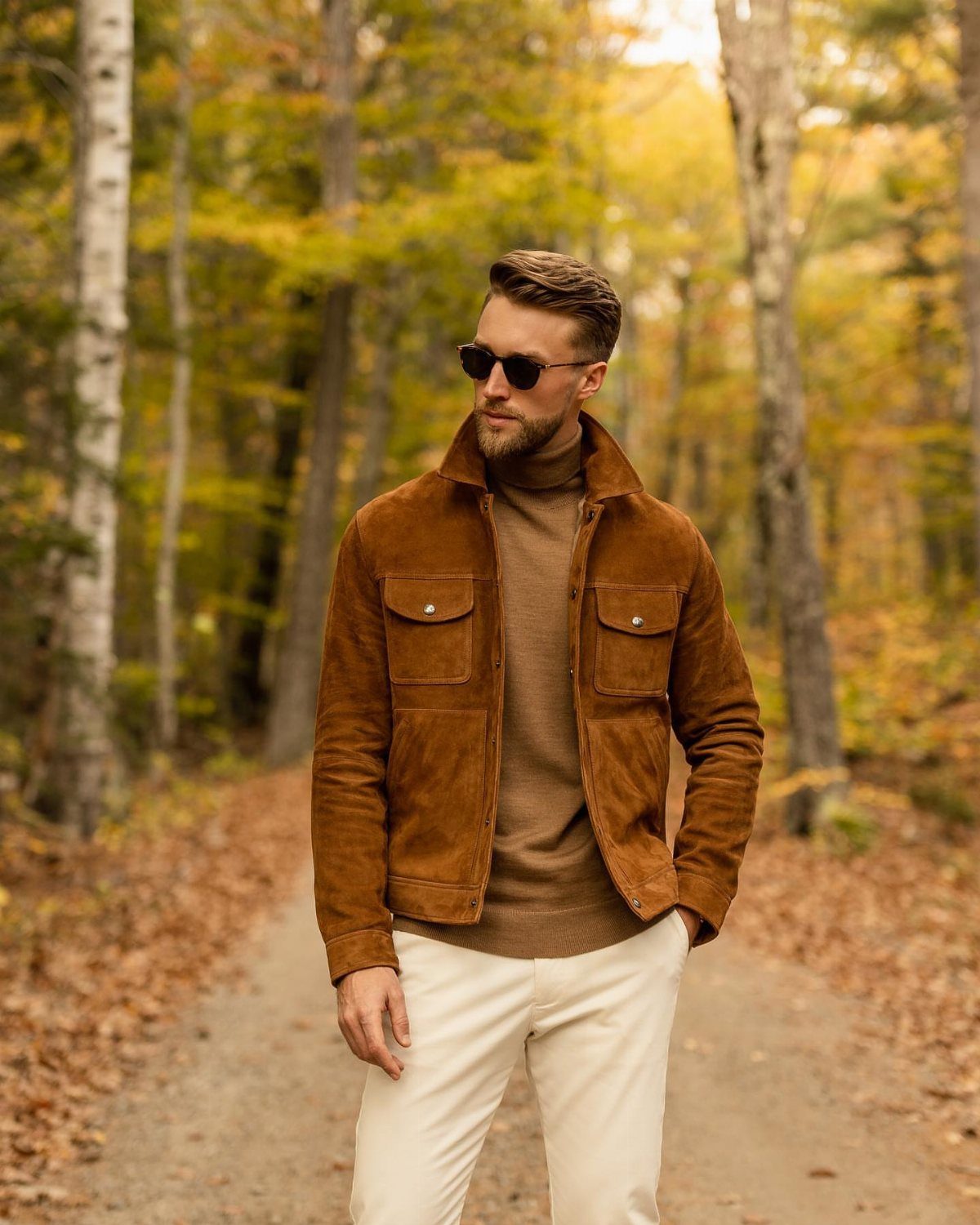 Man in a brown suede jacket and turtleneck sweater standing on a forest path during autumn.